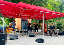 Foto von der Bühne der Band Matterlive! auf einem Kiesplatz am Flussufer der Aare in der Schwellenmätteli. Von links: Gitarrist Nick Perrin, ein nicht genannter Trompeter, Leadsänger Ueli Schmezer, Gebärdensprachdolmetscherin Tanja Joseph, Bassist Michel Poffet, Perkussionist Andy Pupato.