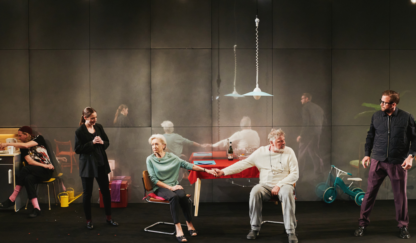 Bühnenfoto mit Ehepaar am Küchentisch, flankiert von weiteren Darstellenden.