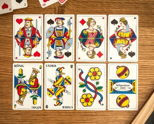 Auf einem Holztisch liegen acht Jasskarten in zwei Reihen. Oben sind französische Bildkarten wie König, Dame und Bube in kräftigen Farben zu sehen. Unten liegen traditionelle Schweizer Jasskarten mit typischen Motiven wie König, Under, einer bunten Blumenkarte und der Rückseite mit dem Schriftzug «Schweizer Jass». Rechts am Rand ist ein Teil eines Holzbretts und zwei rote Bleistifte sichtbar.