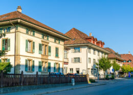 Gebäude der Gemeindeverwaltung von Münchenbuchsee im Jahr 2018. Mehrere traditionelle Verwaltungsgebäude mit Ziegeldach und Fensterläden stehen an einer ruhigen Strasse unter blauem Himmel.