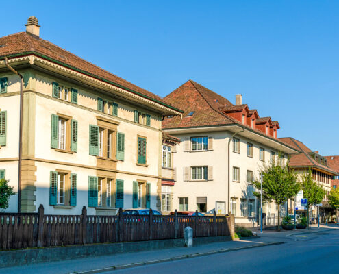 Gebäude der Gemeindeverwaltung von Münchenbuchsee im Jahr 2018. Mehrere traditionelle Verwaltungsgebäude mit Ziegeldach und Fensterläden stehen an einer ruhigen Strasse unter blauem Himmel.
