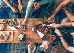 Mehrere Personen sitzen und stehen um einen tiefen Holztisch, auf dem verschiedene Snacks und kleine Speisen wie Brot, Käse, Nüsse, Aufschnitt und Cracker angerichtet sind. Einige Personen halten Gläser mit einem hellen Getränk in der Hand. Die Szene zeigt eine gesellige Tischsituation von oben, mit vielen Händen, die nach Speisen greifen oder Gläser anheben.