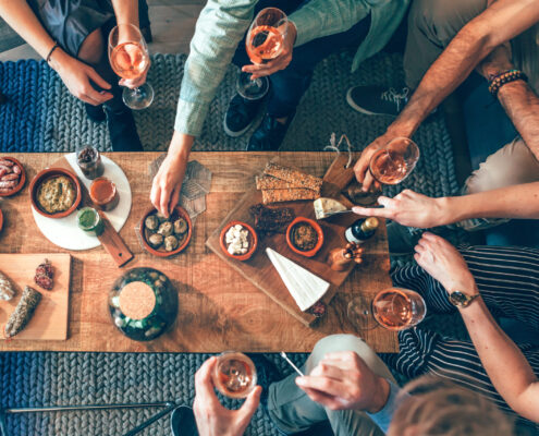 Mehrere Personen sitzen und stehen um einen tiefen Holztisch, auf dem verschiedene Snacks und kleine Speisen wie Brot, Käse, Nüsse, Aufschnitt und Cracker angerichtet sind. Einige Personen halten Gläser mit einem hellen Getränk in der Hand. Die Szene zeigt eine gesellige Tischsituation von oben, mit vielen Händen, die nach Speisen greifen oder Gläser anheben.