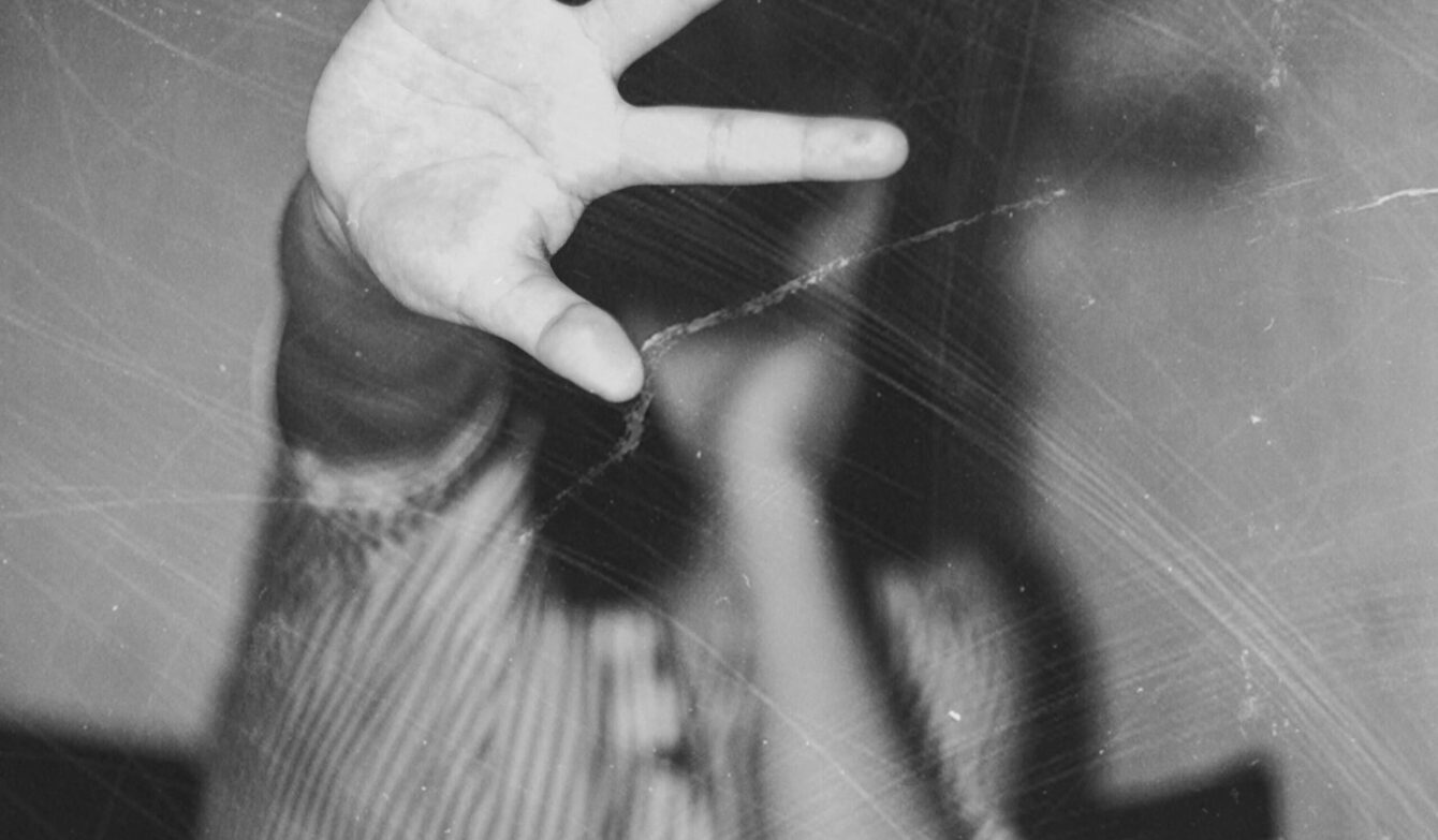 Schwarzweisses Foto in körniger Optik. Im Vordergrund ist eine offene Hand zu sehen, mit gespreizten Fingern, die direkt zur Kamera zeigt und einen grossen Teil des Bildes einnimmt. Die Hand wirkt sehr nah, fast berührend. Im Hintergrund befindet sich eine gebeugte Person, unscharf und teilweise verdeckt durch die erhobene Hand. Der Körper ist zusammengesunken, die Beine sind angezogen, die Knie sichtbar. Die Kleidung zeigt feine Streifen und Punkte. Der Hintergrund ist dunkel und undefiniert, ohne erkennbare Details des Raums. Das Bild vermittelt Distanz und Abwehr durch die nach vorne gerichtete Hand, während der restliche Körper in den Hintergrund zurücktritt.