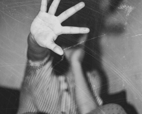 Schwarzweisses Foto in körniger Optik. Im Vordergrund ist eine offene Hand zu sehen, mit gespreizten Fingern, die direkt zur Kamera zeigt und einen grossen Teil des Bildes einnimmt. Die Hand wirkt sehr nah, fast berührend. Im Hintergrund befindet sich eine gebeugte Person, unscharf und teilweise verdeckt durch die erhobene Hand. Der Körper ist zusammengesunken, die Beine sind angezogen, die Knie sichtbar. Die Kleidung zeigt feine Streifen und Punkte. Der Hintergrund ist dunkel und undefiniert, ohne erkennbare Details des Raums. Das Bild vermittelt Distanz und Abwehr durch die nach vorne gerichtete Hand, während der restliche Körper in den Hintergrund zurücktritt.