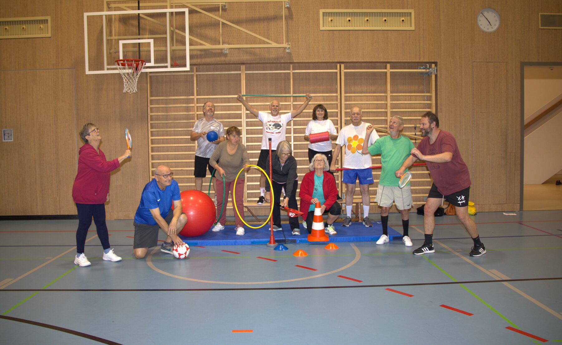 Mehrere Personen befinden sich in einer Turnhalle vor einer Holzwand mit Sprossen und einem Basketballkorb. Die Gruppe ist gemeinsam aktiv und nutzt verschiedene Sport- und Spielgeräte wie Bälle, Reifen, Kegel und Schläger. Einige Personen stehen, andere sitzen oder knien auf Matten. Die Szene wirkt wie ein gemeinsames Bewegungs- oder Trainingsangebot, bei dem unterschiedliche Übungen gleichzeitig stattfinden.