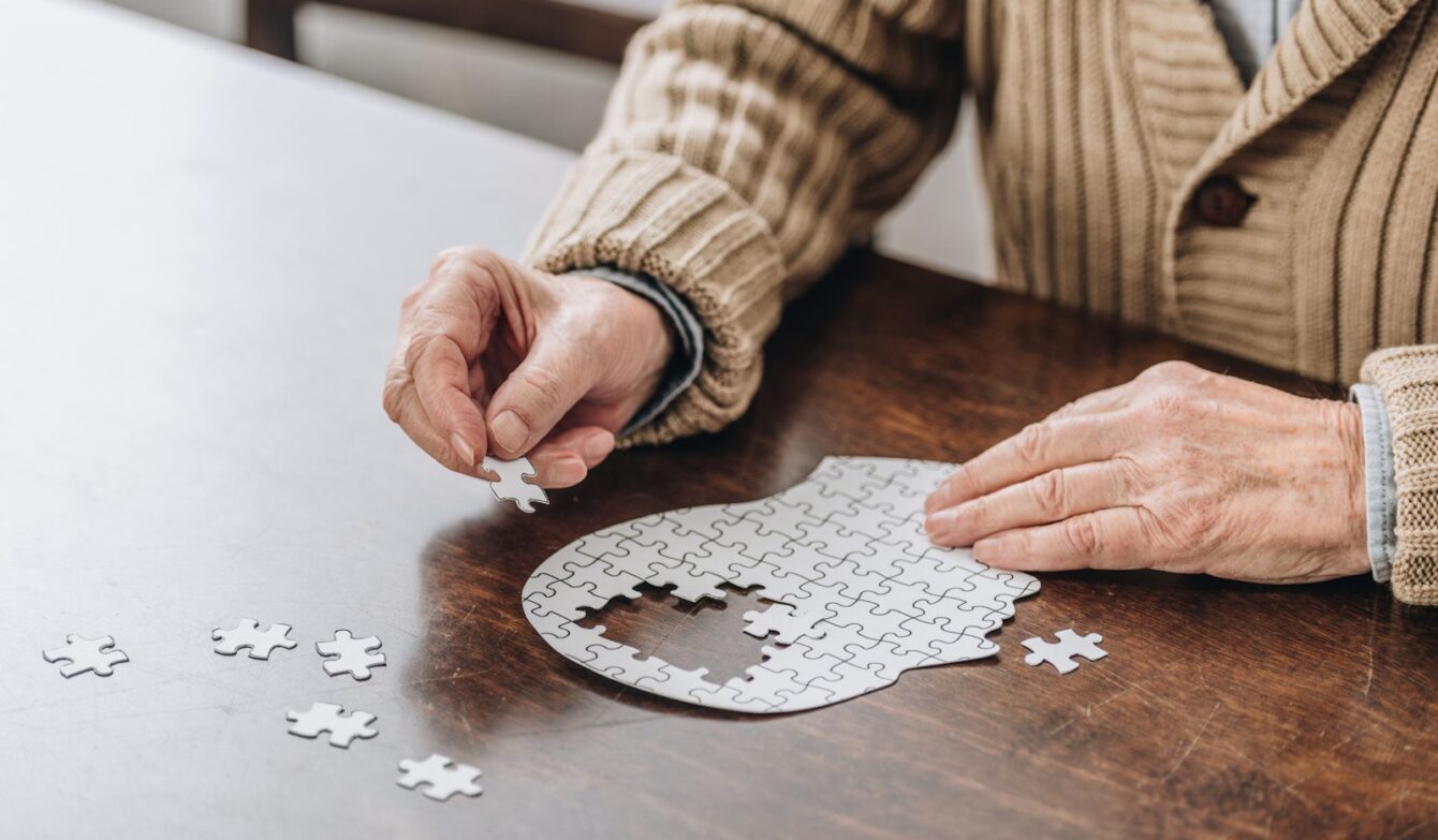 Eine Person sitzt an einem Tisch und setzt ein Puzzle zusammen, das die Form eines menschlichen Kopfes hat. Viele Teile liegen bereits an ihrem Platz, aber in der Mitte fehlt noch ein Stück. Die Hände halten ein kleines Puzzleteil, das gleich eingesetzt werden soll. Weitere lose Teile liegen auf dem Tisch daneben.