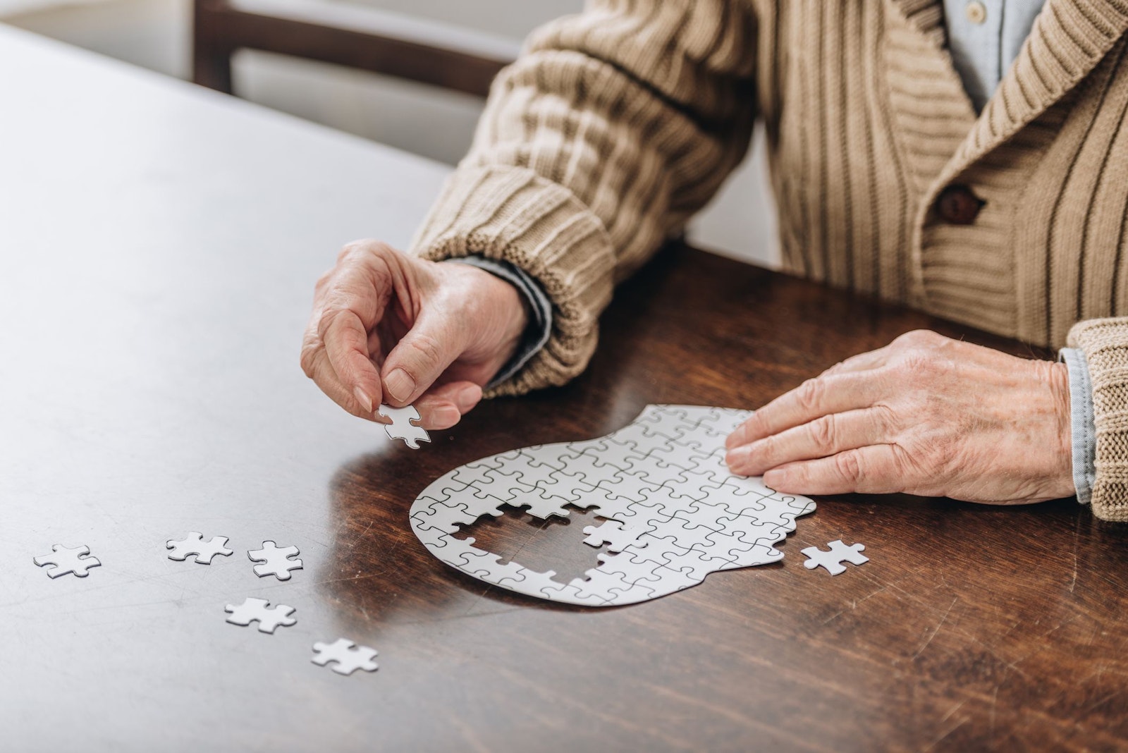 Eine Person sitzt an einem Tisch und setzt ein Puzzle zusammen, das die Form eines menschlichen Kopfes hat. Viele Teile liegen bereits an ihrem Platz, aber in der Mitte fehlt noch ein Stück. Die Hände halten ein kleines Puzzleteil, das gleich eingesetzt werden soll. Weitere lose Teile liegen auf dem Tisch daneben.